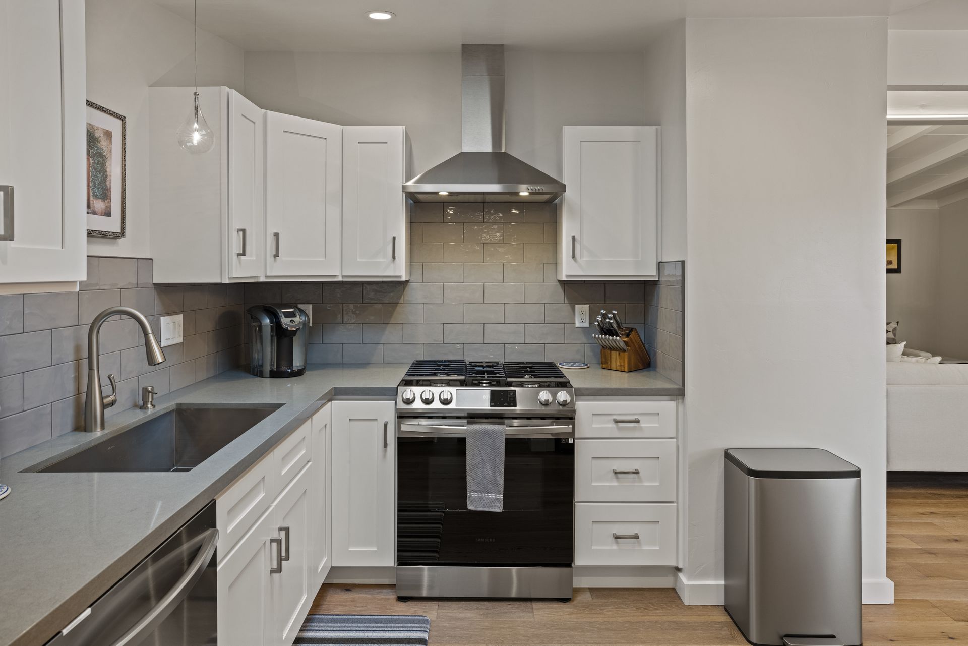 A kitchen with white cabinets , a stove , a sink , and a trash can.