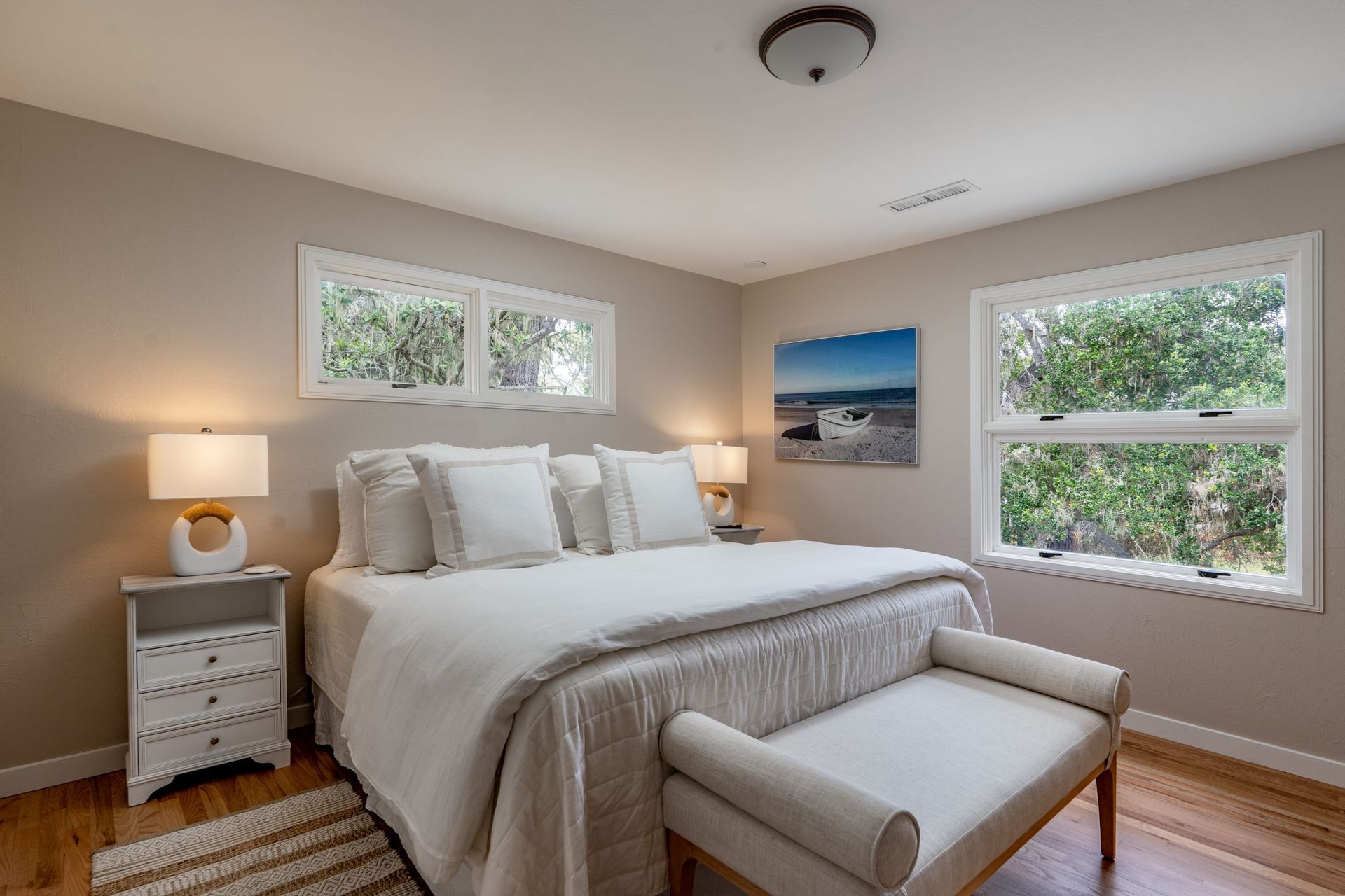 A bedroom with a large bed , a bench , lamps and two windows.