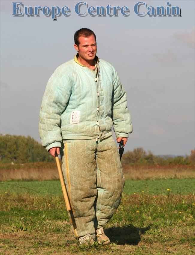 Un homme en combinaison de protection, tenant un bâton, se tient dans un champ. Le logo « Europe Centre Canin » est visible en haut.