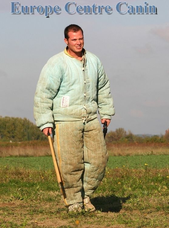Homme en combinaison de protection rembourrée, tenant un bâton, debout dans un champ. Logo « Europe Centre Canin » en haut.