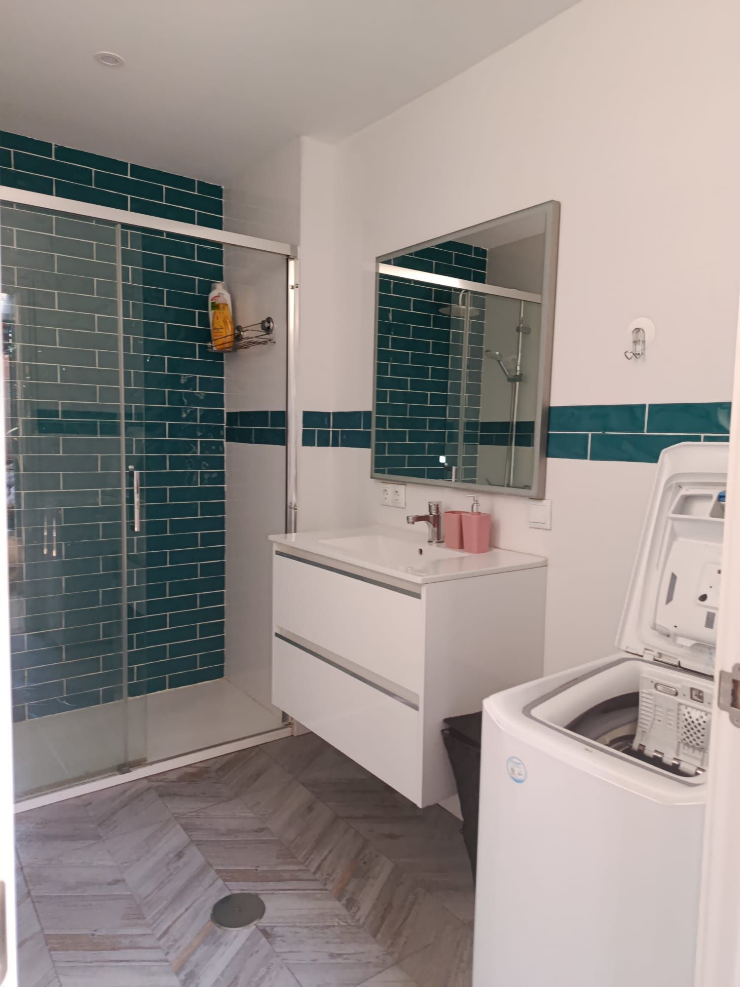 Baño moderno con ducha de azulejos verde azulado, lavabo blanco, espejo y lavadora.