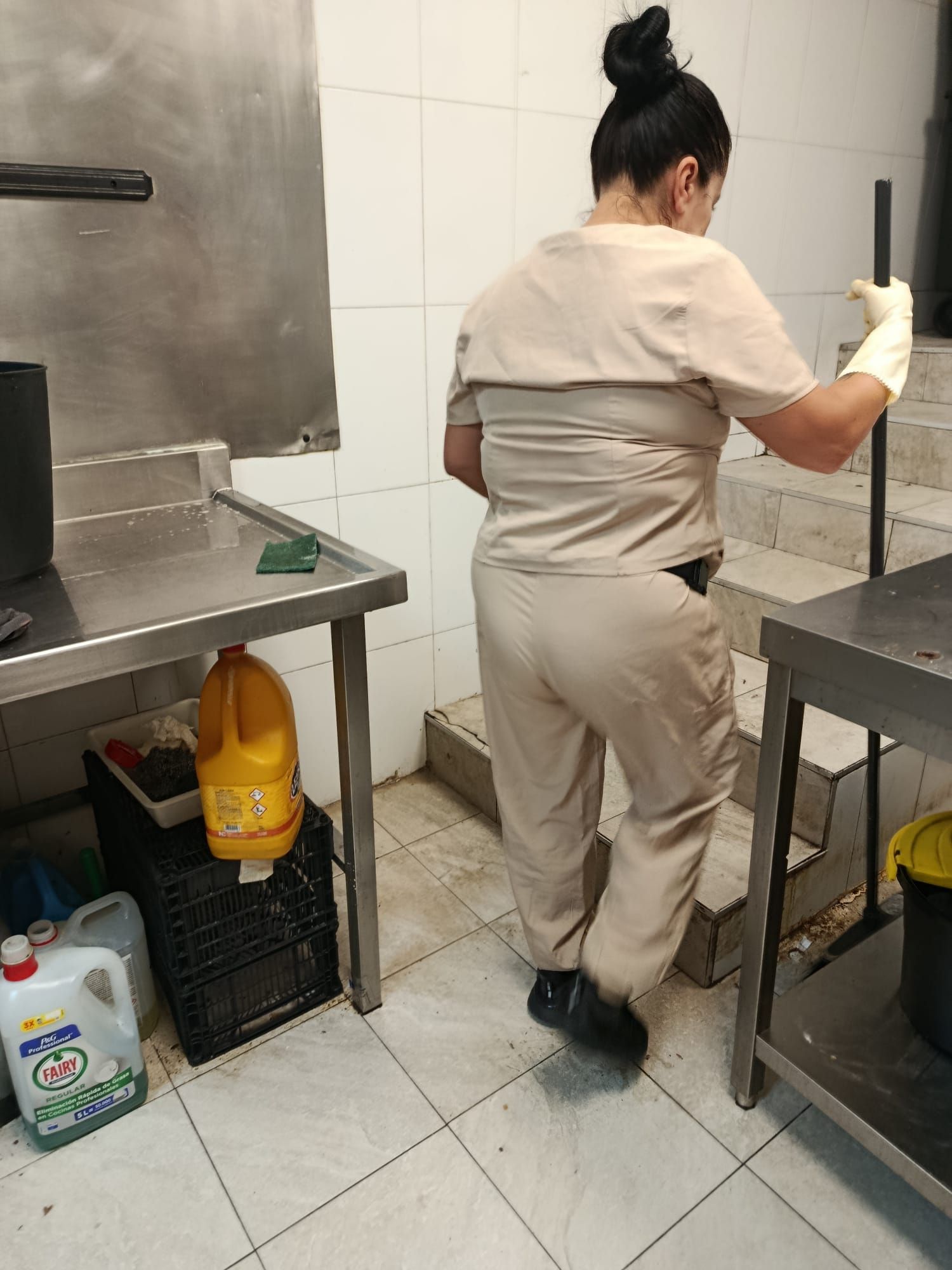 Persona con uniforme beige trabajando en una cocina alicatada junto a encimeras de acero inoxidable.