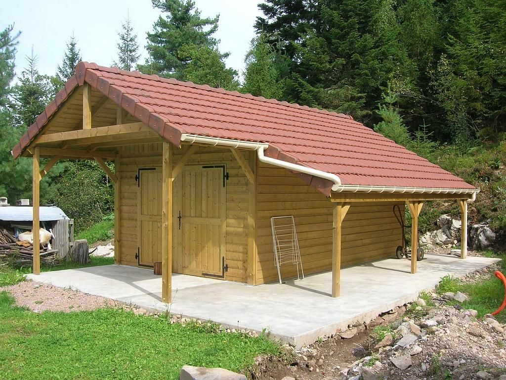 Un abri en rondins de bois avec un toit de tuiles rouges et une terrasse en béton, niché dans une clairière herbeuse bordée d'arbres.