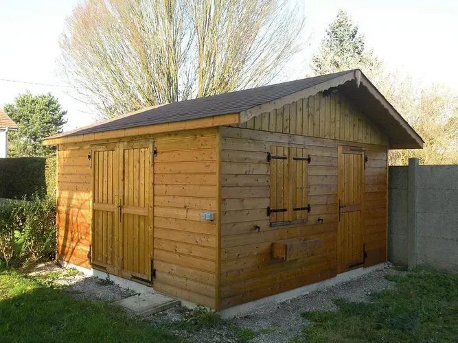Un abri en bois avec double porte, une fenêtre à volets et un toit à pignon, situé sur une base en béton dans une cour gazonnée.