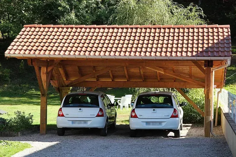 Deux voitures blanches garées côte à côte sous un abri en bois au toit de tuiles rouges, sur une allée de gravier.