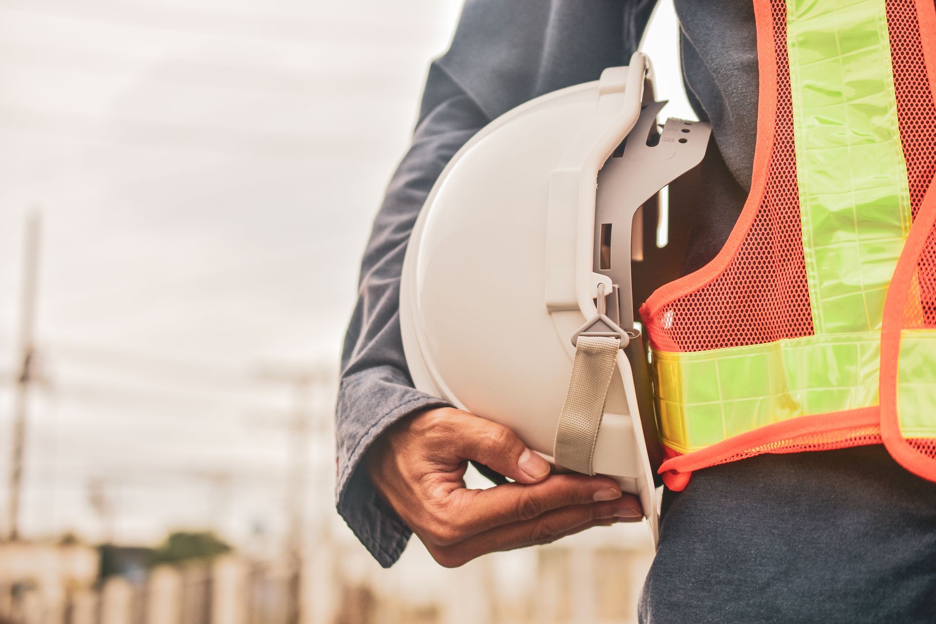 Une personne portant un gilet de sécurité haute visibilité et tenant un casque de chantier blanc à ses côtés, sur fond industriel.