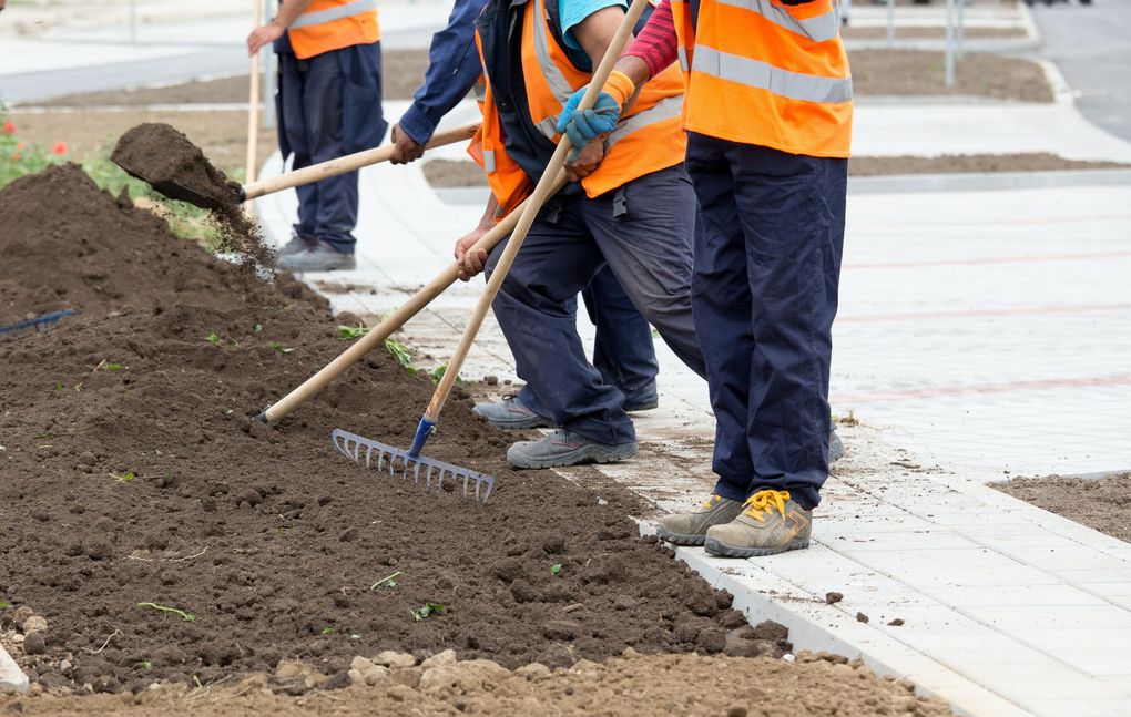 Des ouvriers vêtus de gilets de sécurité orange utilisent des râteaux et des pelles pour étaler de la terre dans un espace paysager extérieur.