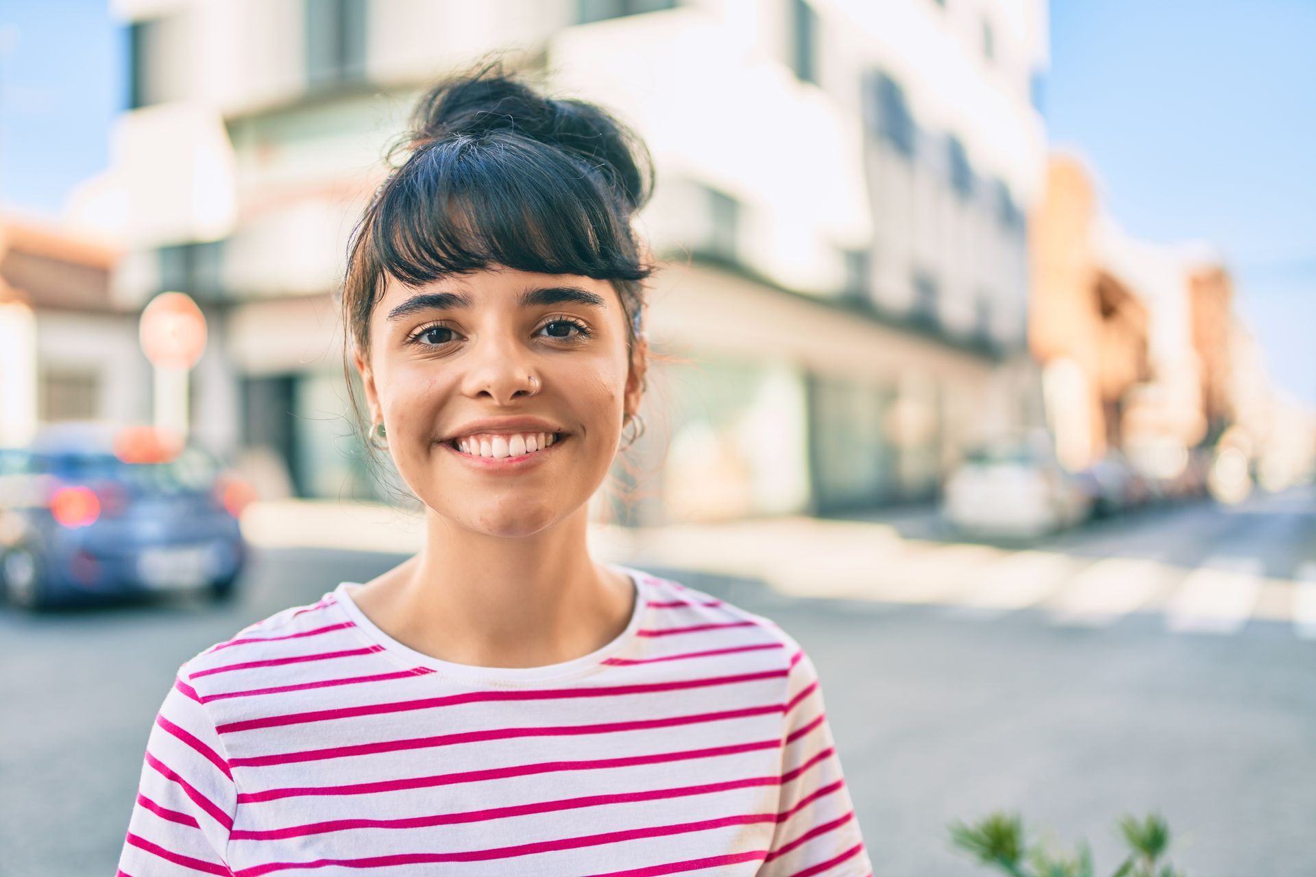 Une personne souriante aux cheveux foncés coiffés en chignon porte une chemise à rayures roses et blanches à l'extérieur, dans une rue floue de la ville.