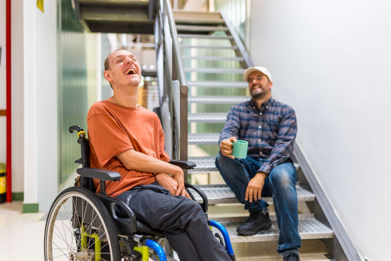 Une personne en fauteuil roulant rit tandis qu'une autre personne, assise sur un escalier voisin, tient une tasse et sourit.