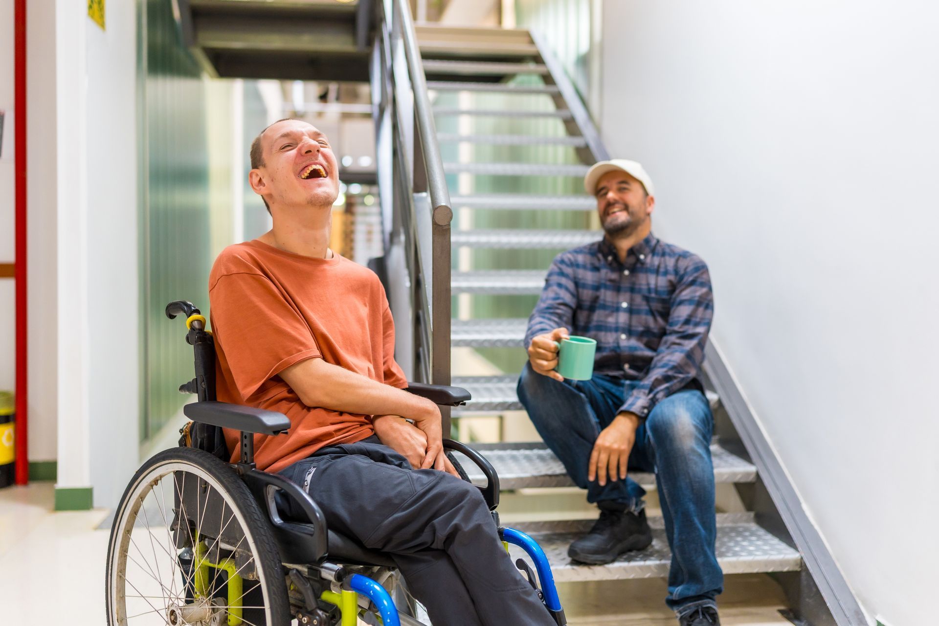 Une personne en fauteuil roulant rit tandis qu'une autre personne, assise sur un escalier voisin, tient une tasse et sourit.