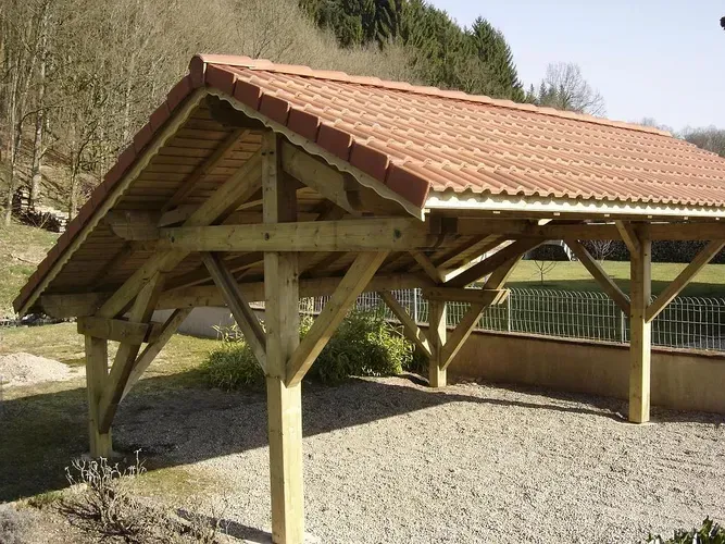 Un abri de voiture en bois, ouvert sur les côtés, avec un toit à pignon en tuiles de terre cuite, posé sur une surface de gravier.