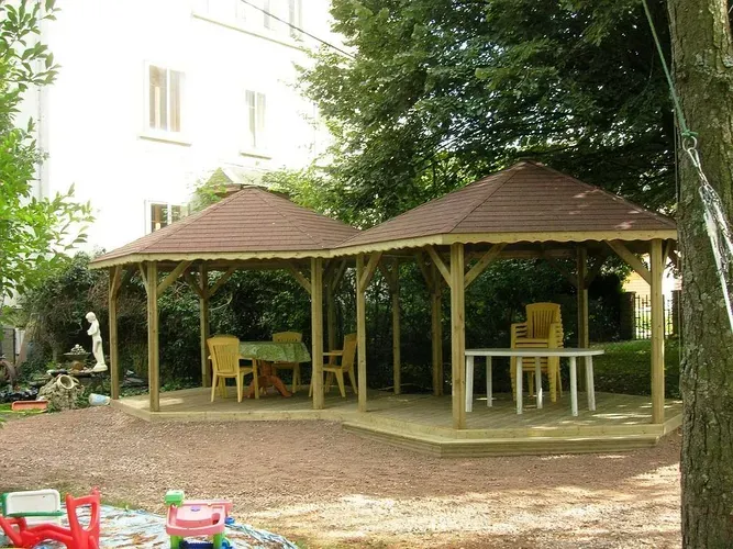 Deux kiosques en bois reliés entre eux, aux toits bruns, se dressent sur une terrasse de gravier dans un jardin ; chacun est meublé d'une table et de chaises.