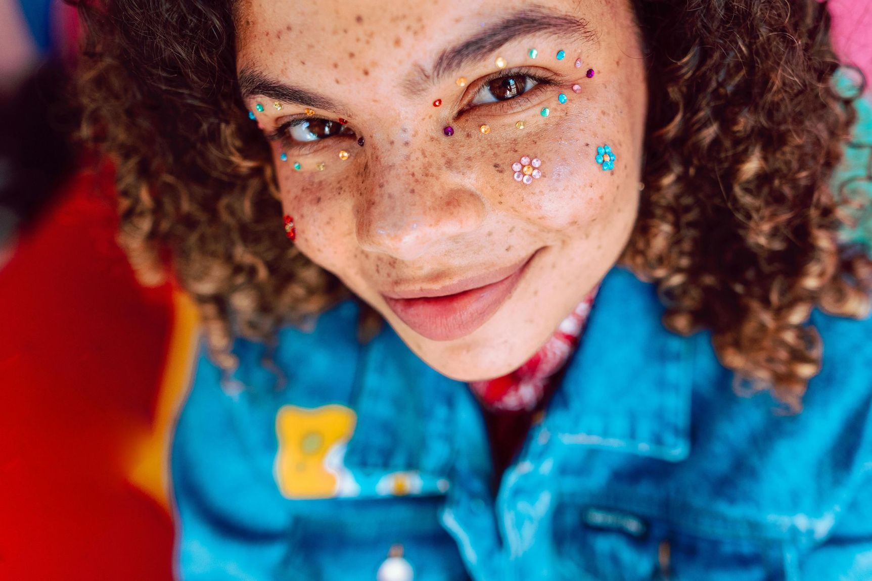 Une personne aux cheveux bouclés et au visage orné de taches de rousseur et de petits bijoux colorés, portant une veste en jean bleu vif.