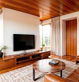Sala de estar moderna con techo de madera, televisor montado en la pared, mesa de centro redonda y ventana grande con cortinas.