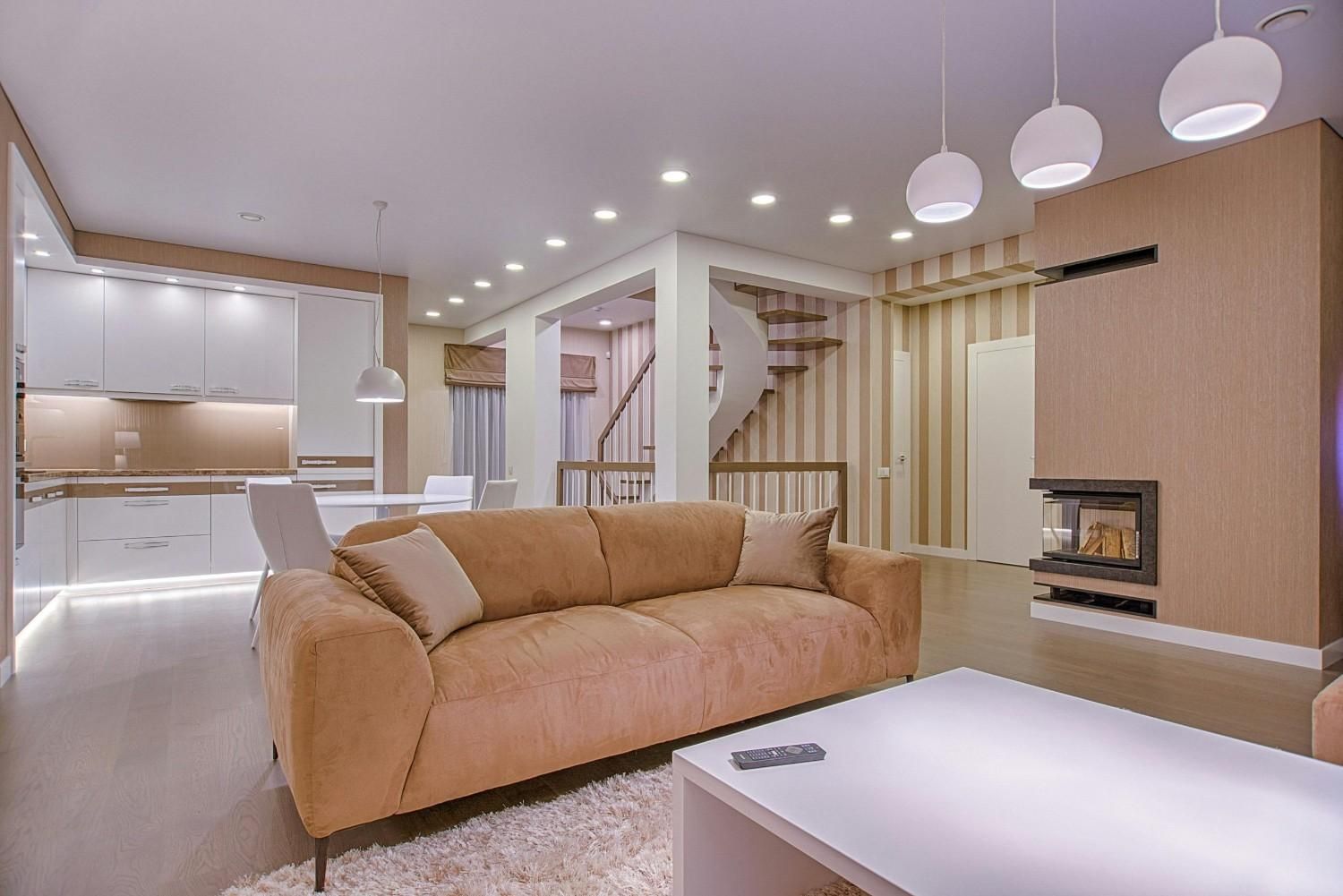 Sala de estar moderna de concepto abierto con sofá color canela, mesa de centro blanca, lámparas colgantes y escalera al fondo.