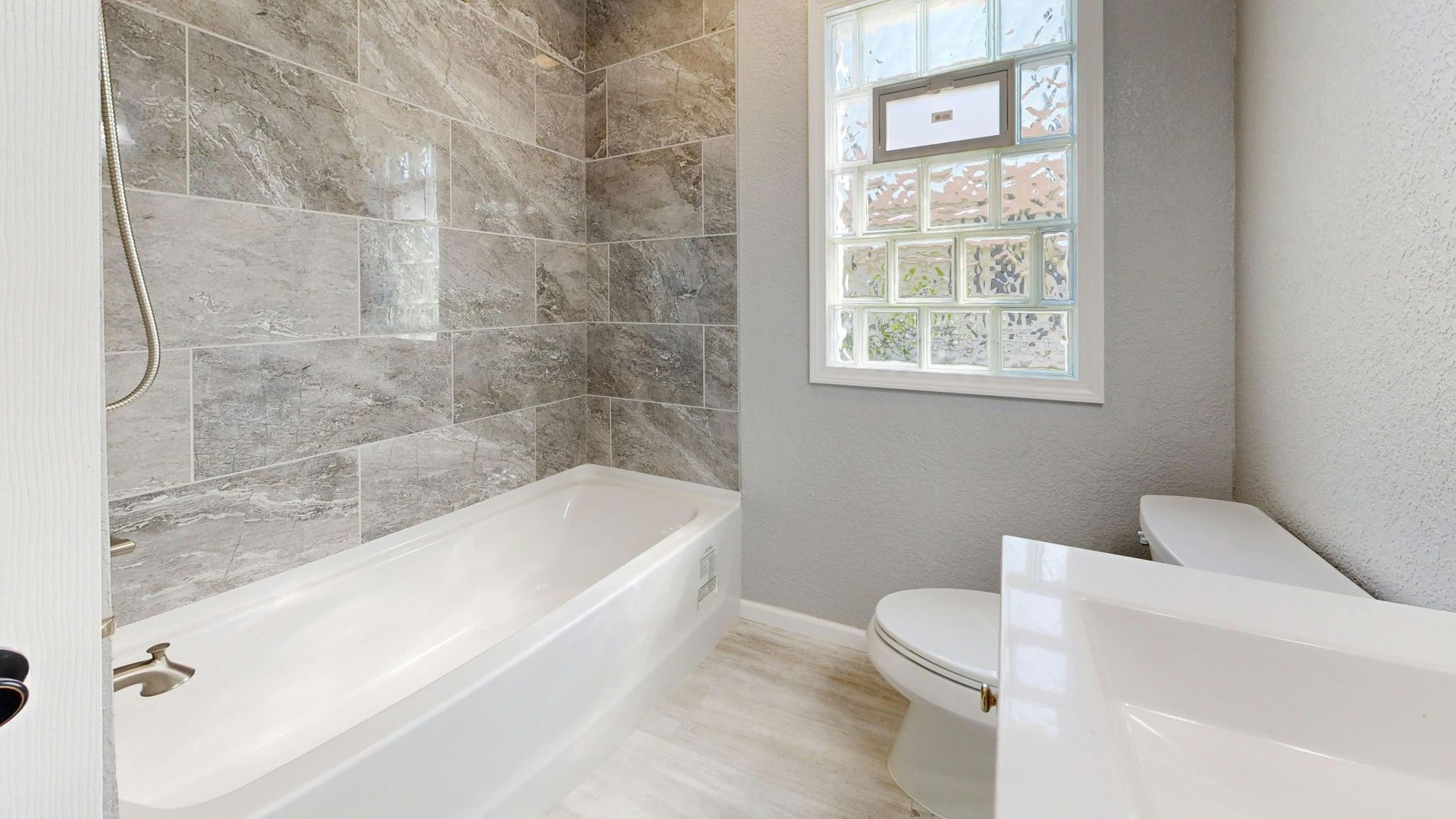 Baño luminoso con bañera blanca, pared de ducha de piedra gris y ventana sobre el inodoro.
