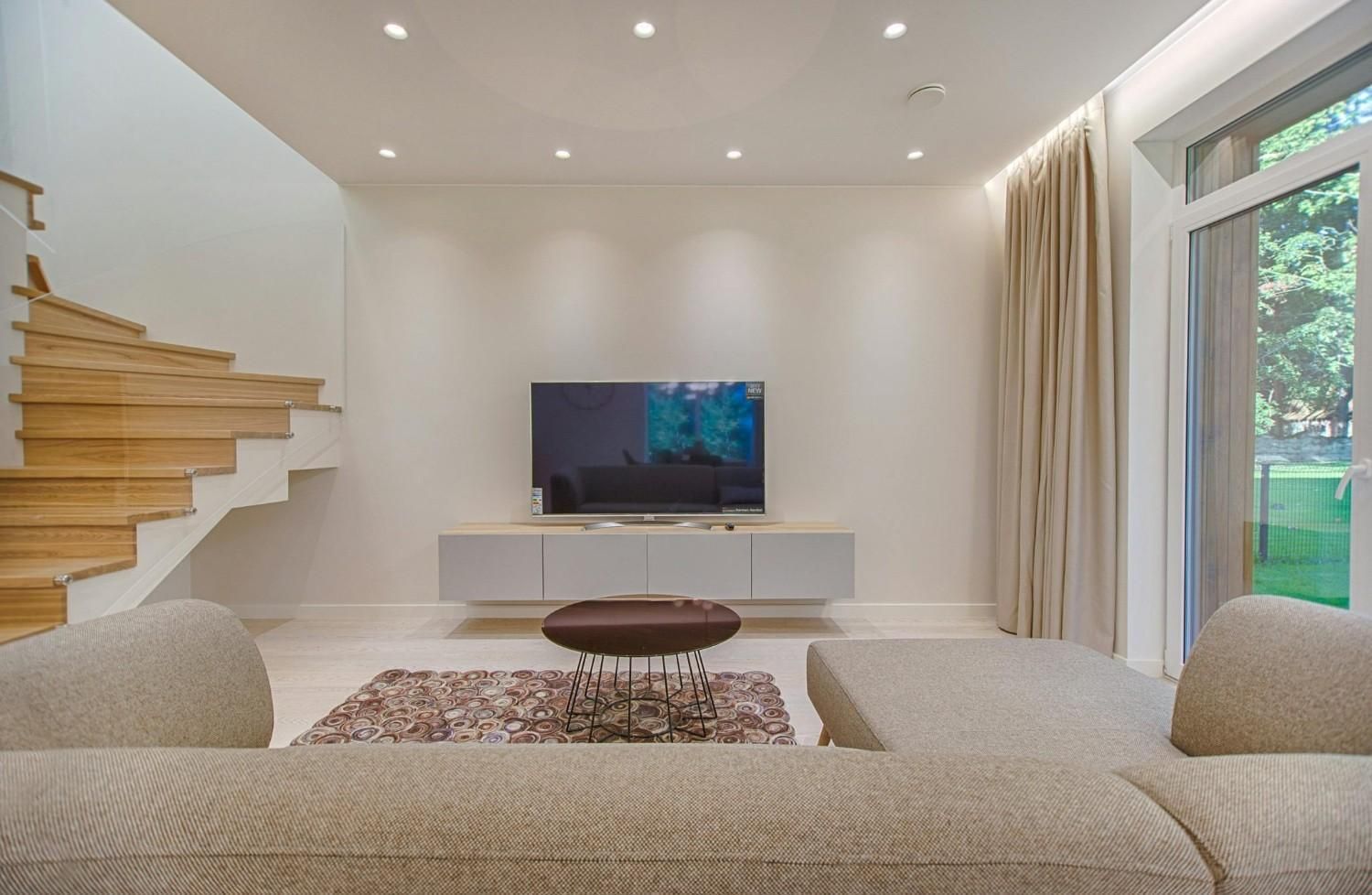 Sala de estar moderna con sofá modular, televisor montado en la pared, escalera de madera y gran ventanal.