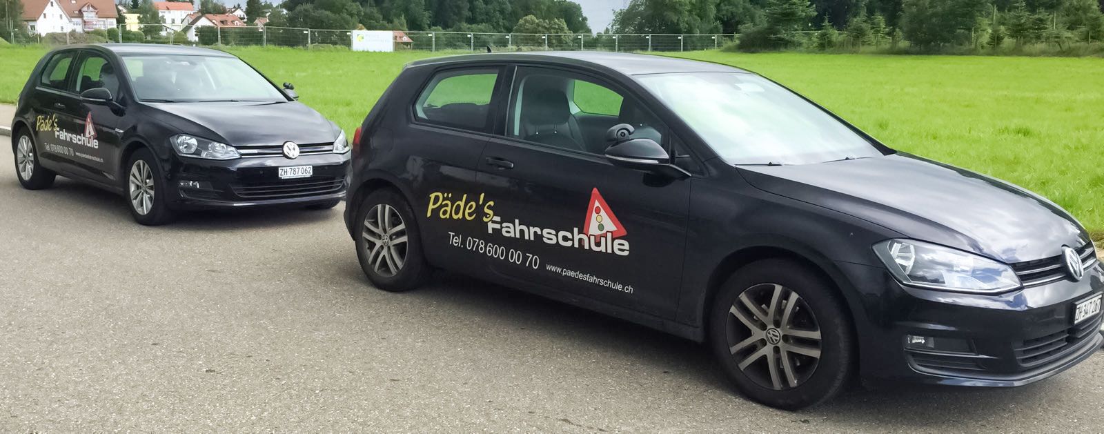 Zwei schwarze Volkswagen Golf Fahrlehrerautos auf einer Asphaltstraße mit einer grünen Wiese und Häusern im Hintergrund.