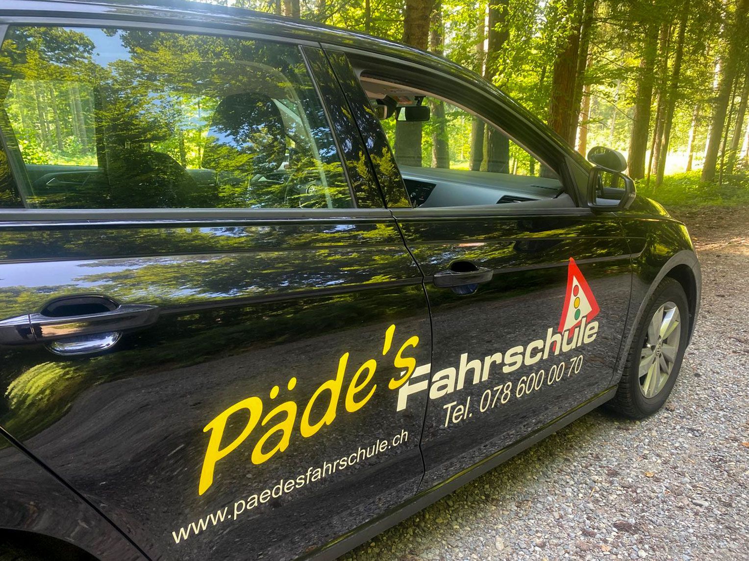 Schwarzes Auto mit „Päde’s Fahrschule“-Logo und Fahrschulschild; im Freien.