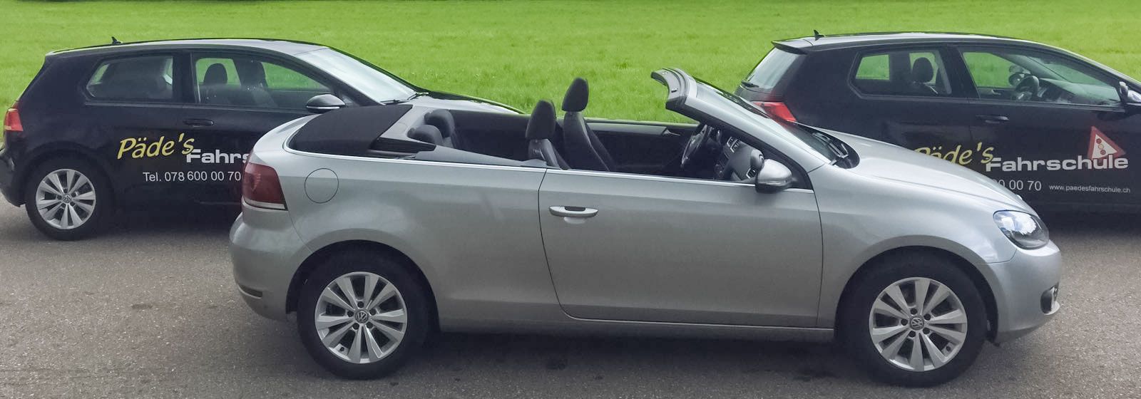 Silbernes Cabrio, flankiert von zwei schwarzen Autos auf einer gepflasterten Fläche, mit grünem Gras im Hintergrund.