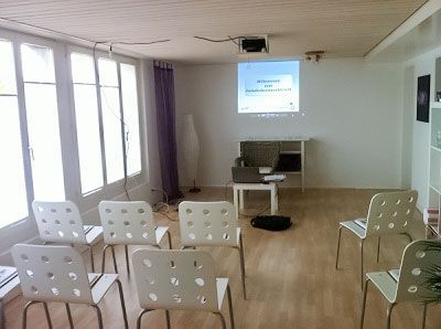 Leerer Klassenraum mit weißen Stühlen vor einem Bildschirm, auf dem eine Präsentation läuft. Sonnenlicht fällt durch die Fenster.