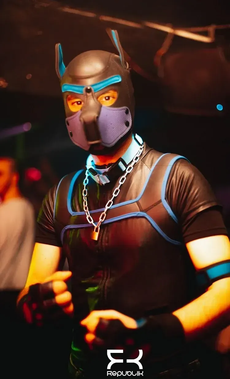 Persona con máscara de cachorro y arnés en un club; ropa oscura, detalles en azul, cadena plateada, manos cerca de la cintura.