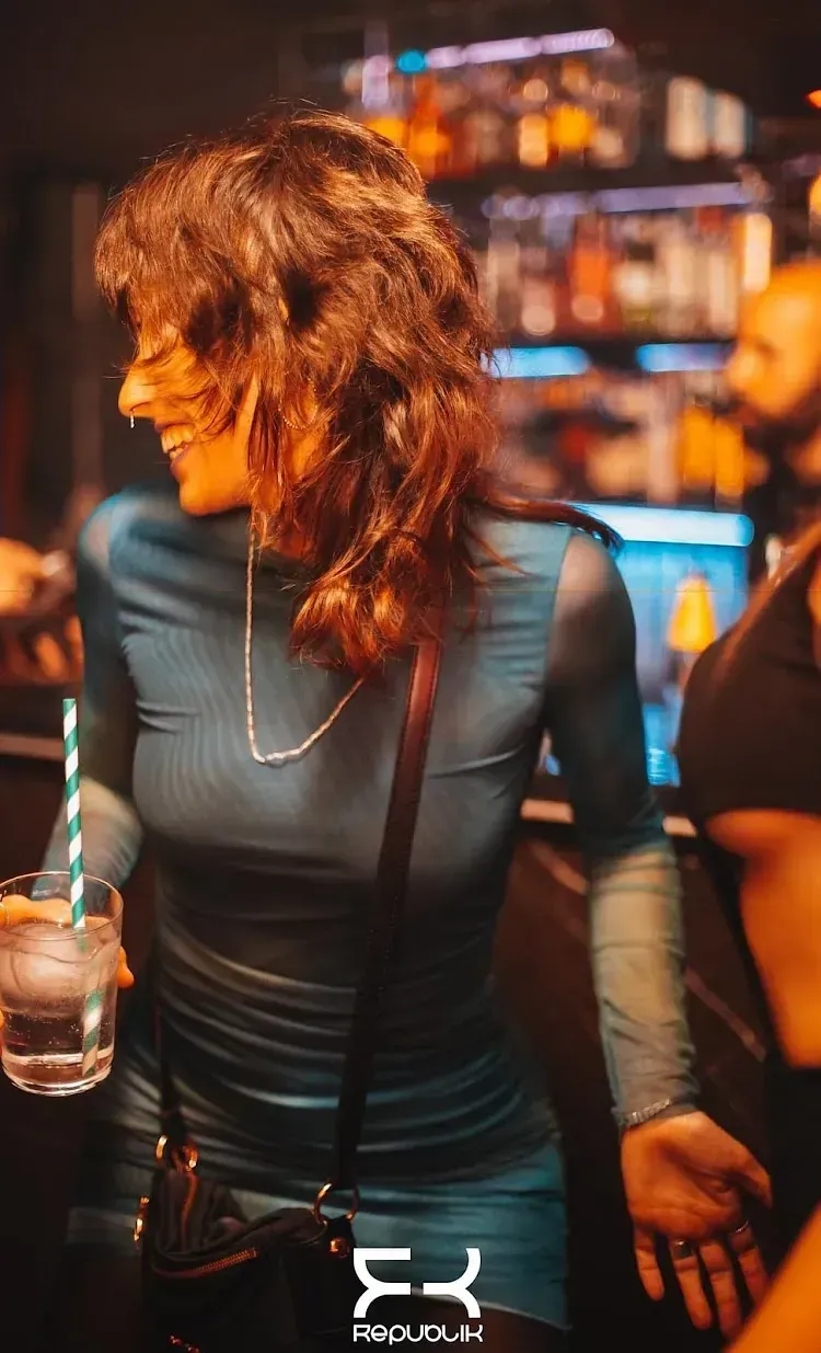 Una mujer con un vestido azul sonríe, sosteniendo una bebida, en un bar.
