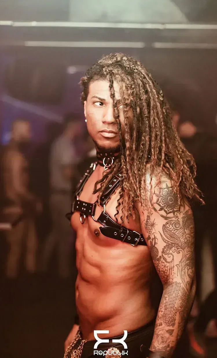 Hombre con rastas, tatuajes y arnés de cuero posa en un club.