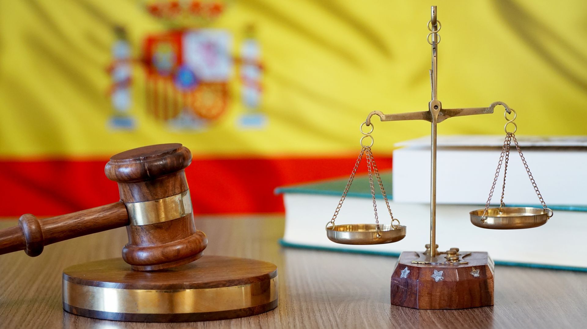 Un mazo de juez y una balanza de la justicia están sobre una mesa de madera delante de una bandera española.