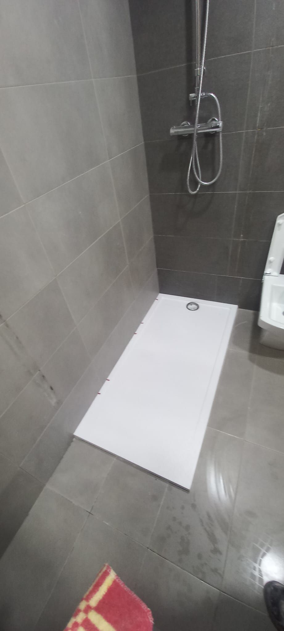 Un plato de ducha rectangular blanco descansa sobre un suelo de baldosas en un baño con azulejos grises en las paredes