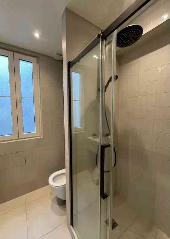 Un baño moderno con paredes de azulejos de color claro, una ducha acristalada y un inodoro blanco junto a una ventana.