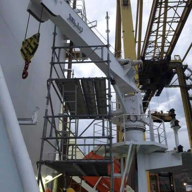 Grúa con andamio fijado a un barco; gancho amarillo, estructura blanca y gris, entorno industrial.