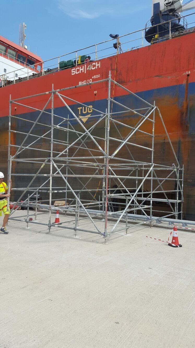 Andamio instalado al lado de un gran barco rojo, y un trabajador con equipo amarillo se encuentra cerca.