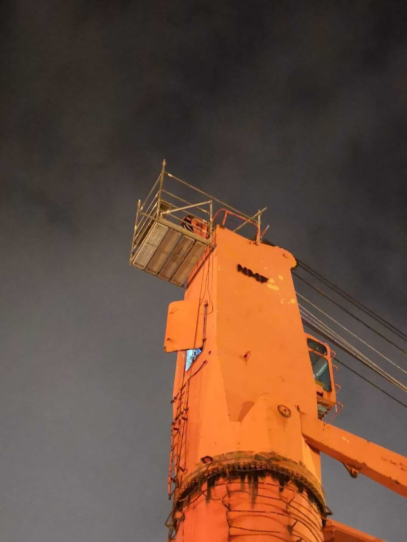 Grúa industrial naranja contra un cielo oscuro, con una plataforma de andamio en la parte superior.