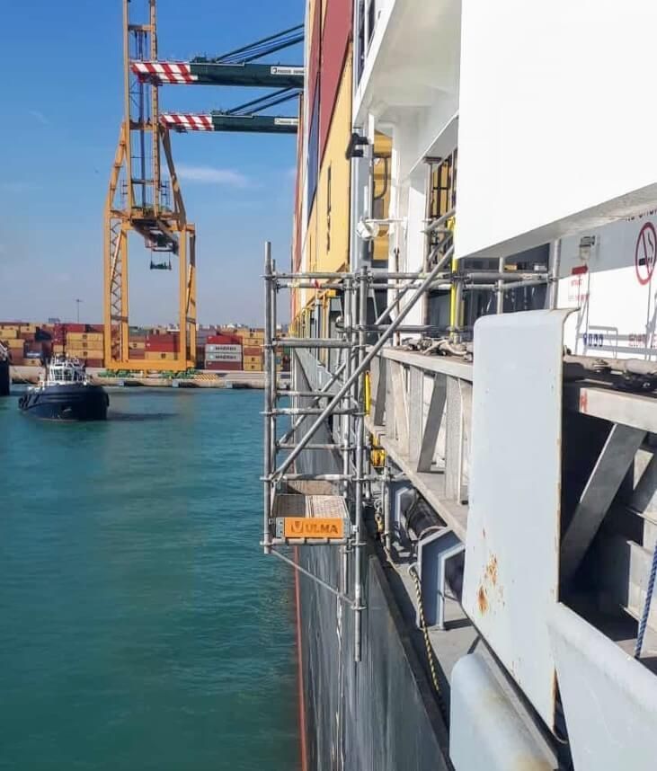 Andamios junto a un barco de carga en un puerto, con grúas y remolcador al fondo.