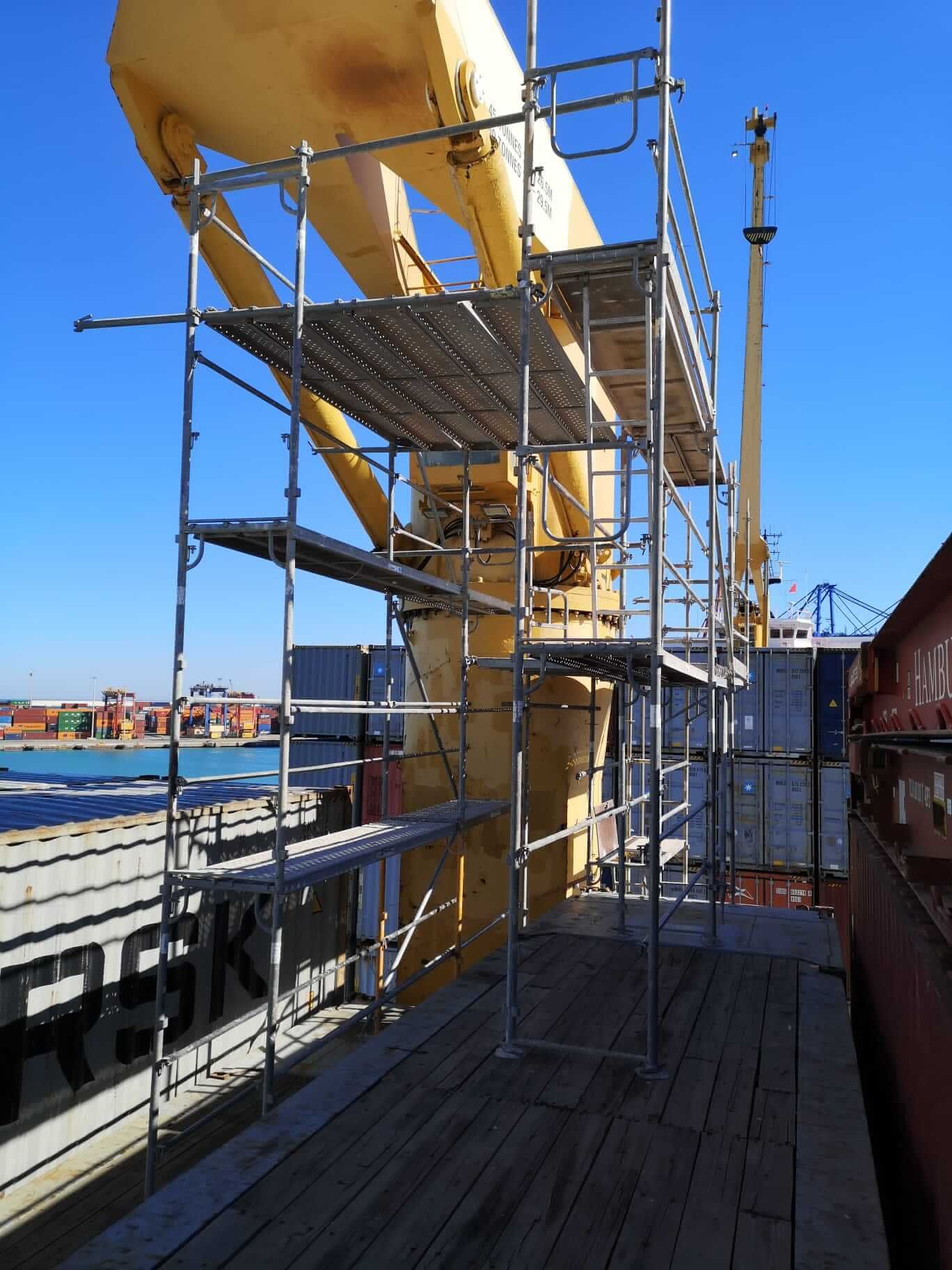 Andamio construido junto a una grúa amarilla en un muelle con contenedores de carga, cielo azul.