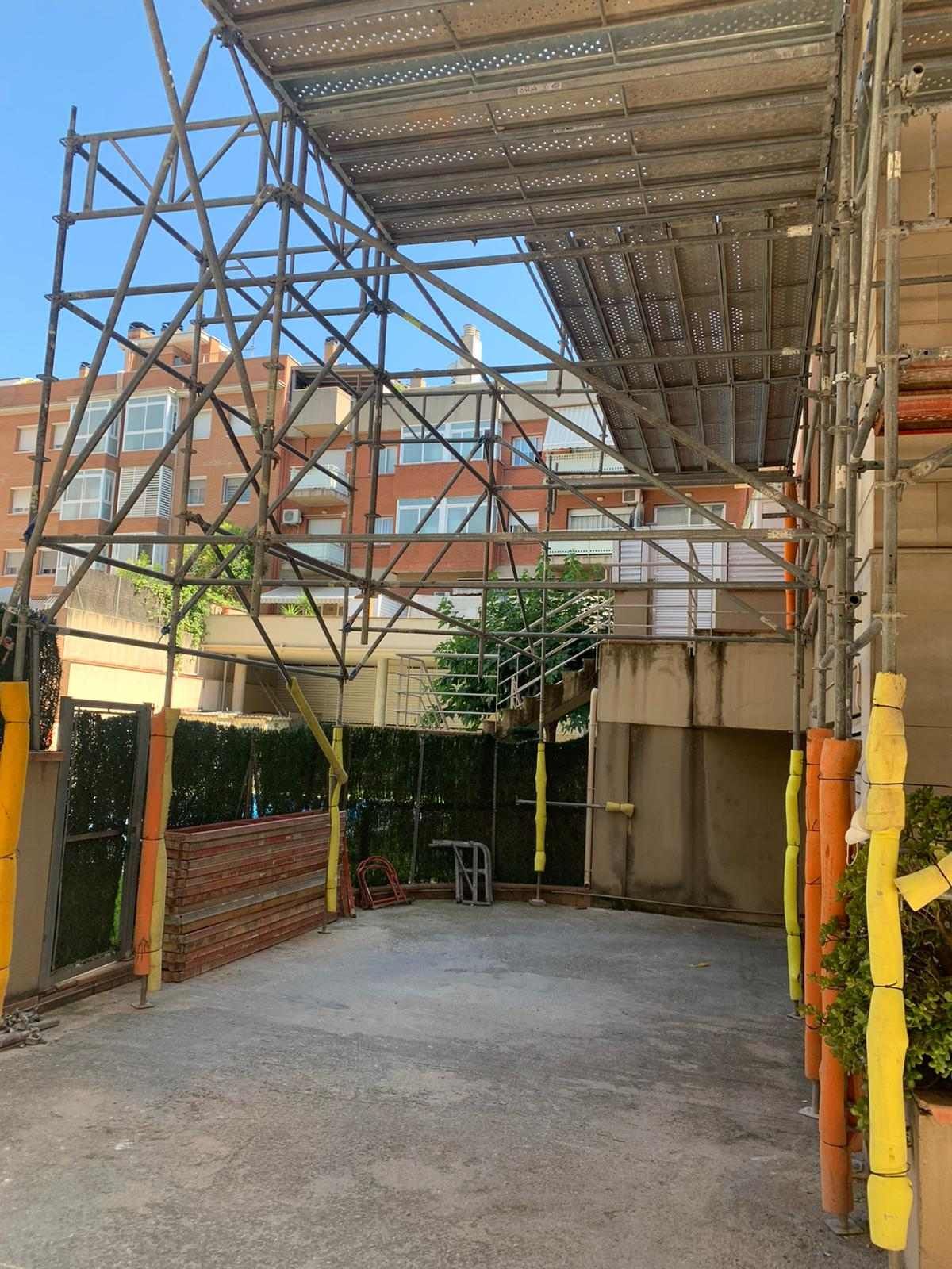 Un andamio cubre un estrecho pasillo exterior. Se ven edificios y vegetación.