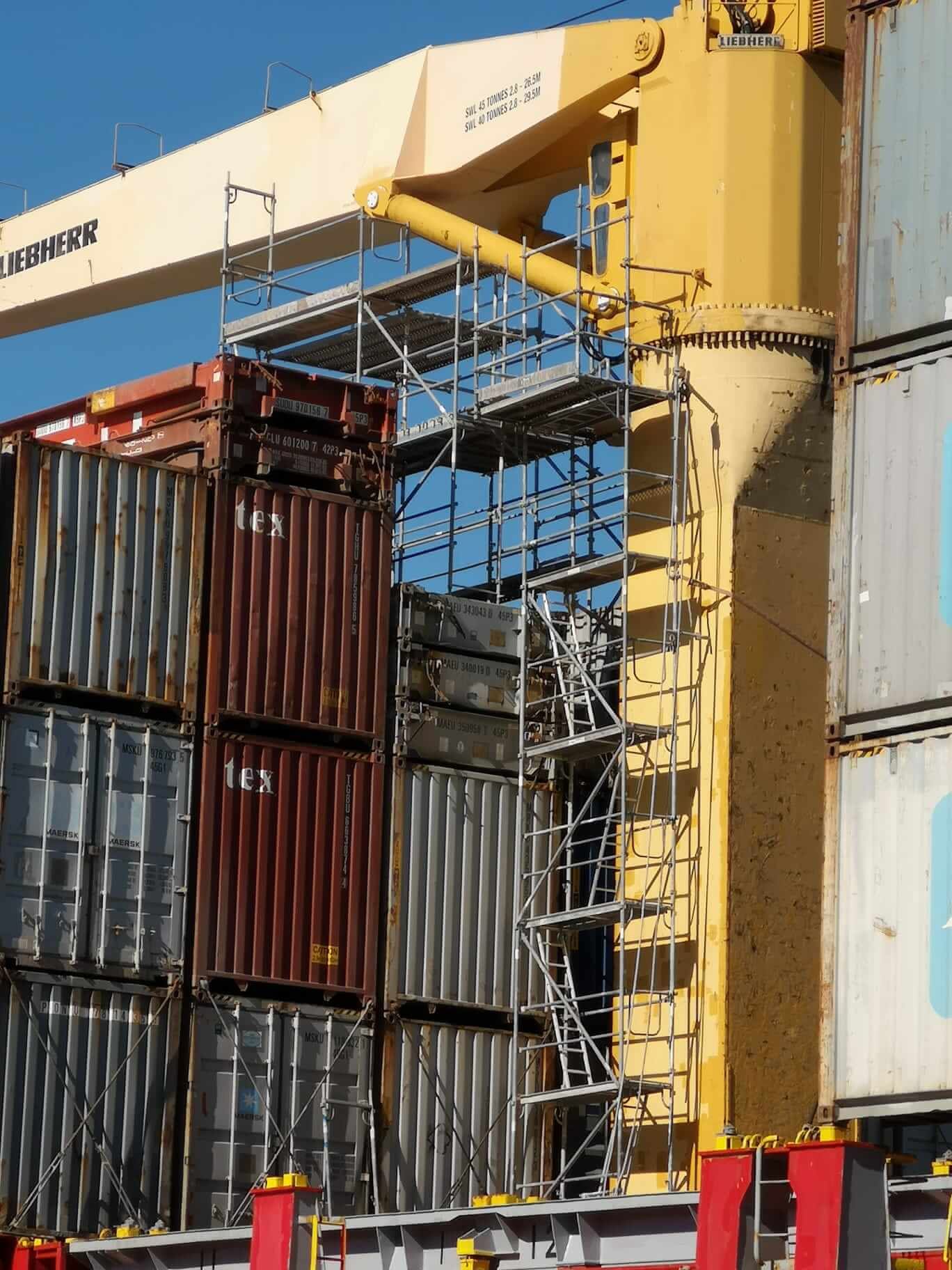 Grúa amarilla con andamios acoplados junto a contenedores de envío apilados en un puerto.