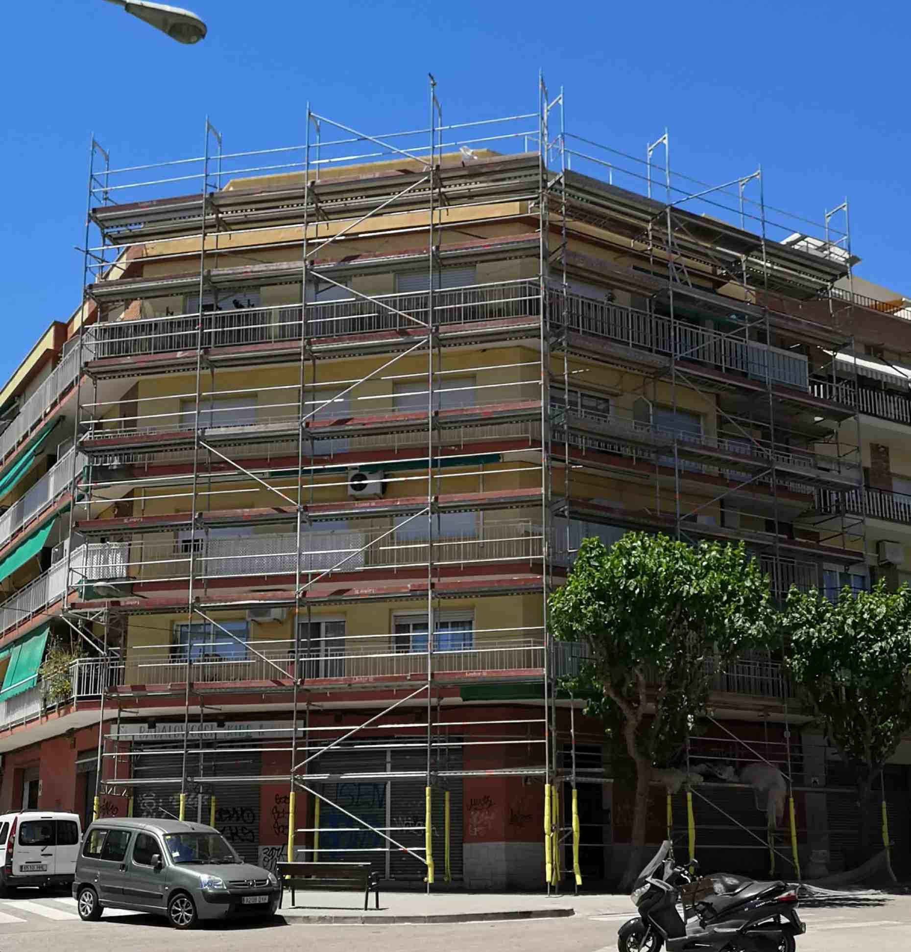 Edificio en remodelación. Andamios rodean el exterior. Fachada amarilla, escena callejera, autos estacionados.