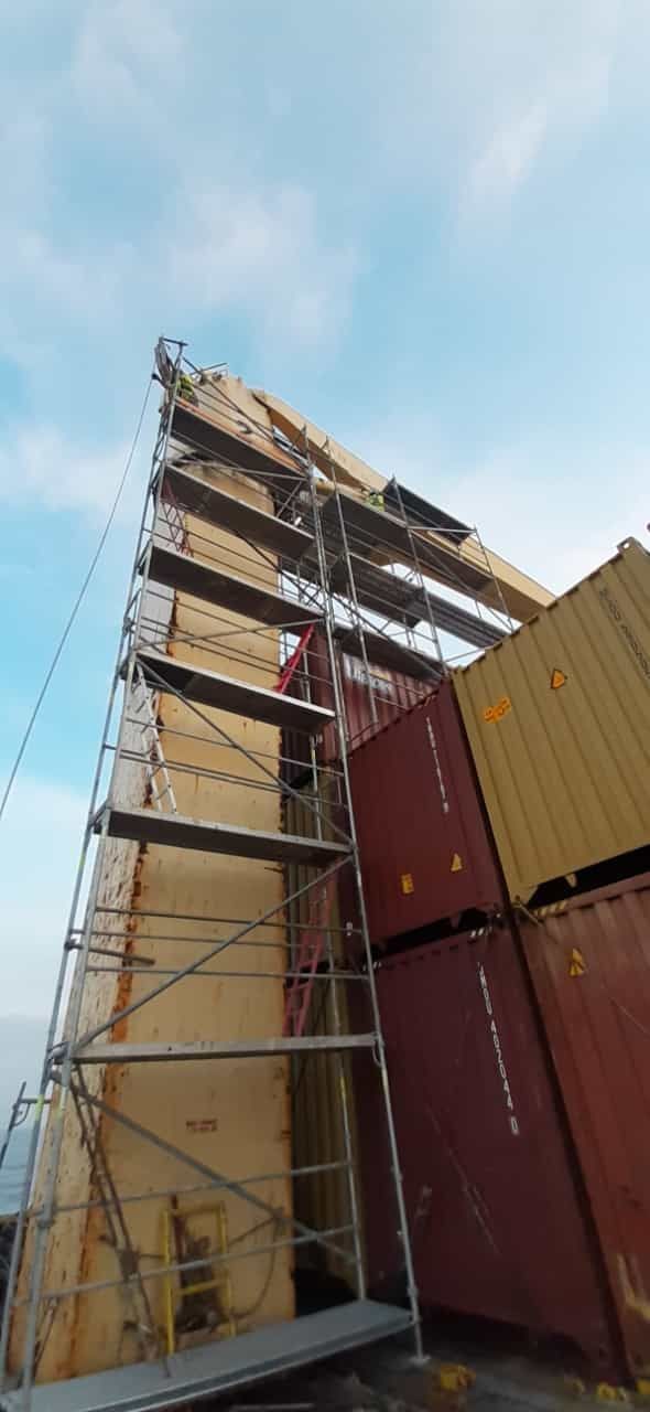 Andamios en un barco con contenedores apilados. Cielo azul al fondo.