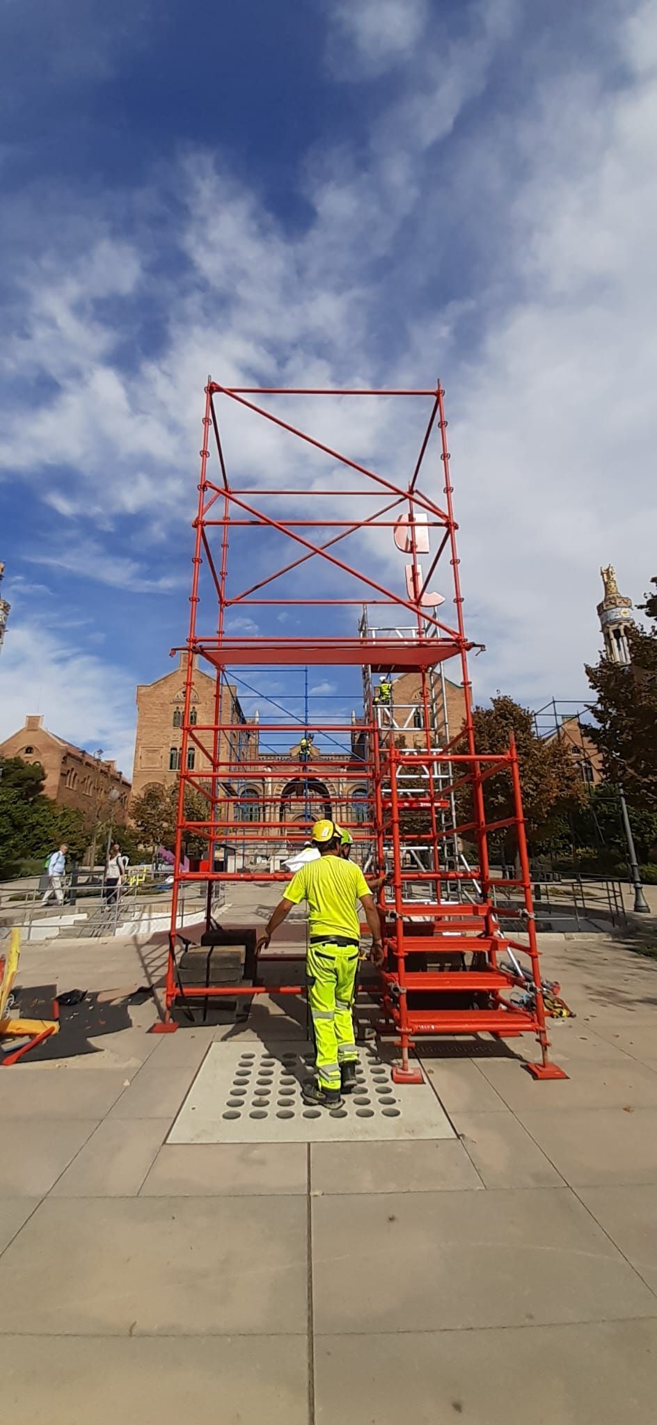 Una persona con equipo de seguridad amarillo camina hacia un andamio rojo sobre hormigón, bajo un cielo azul.