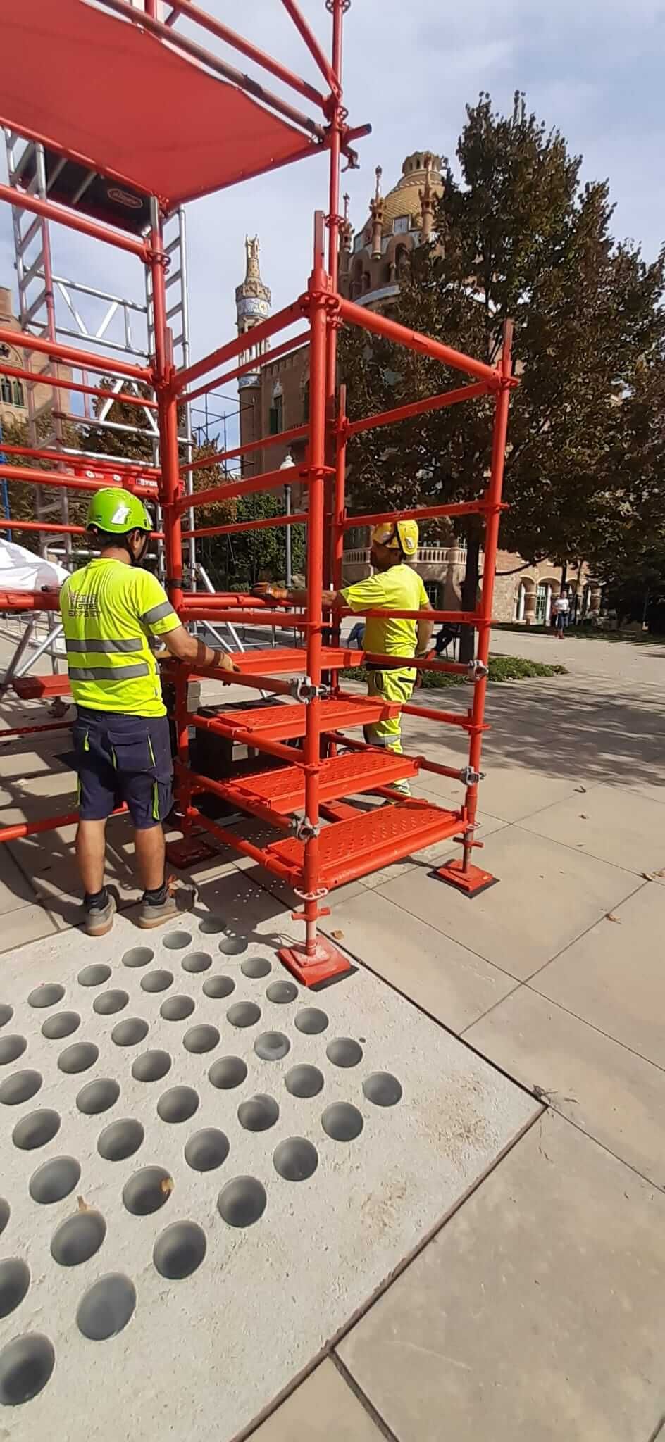 Dos trabajadores de la construcción con chalecos de neón montan andamios de color naranja sobre una superficie de hormigón.