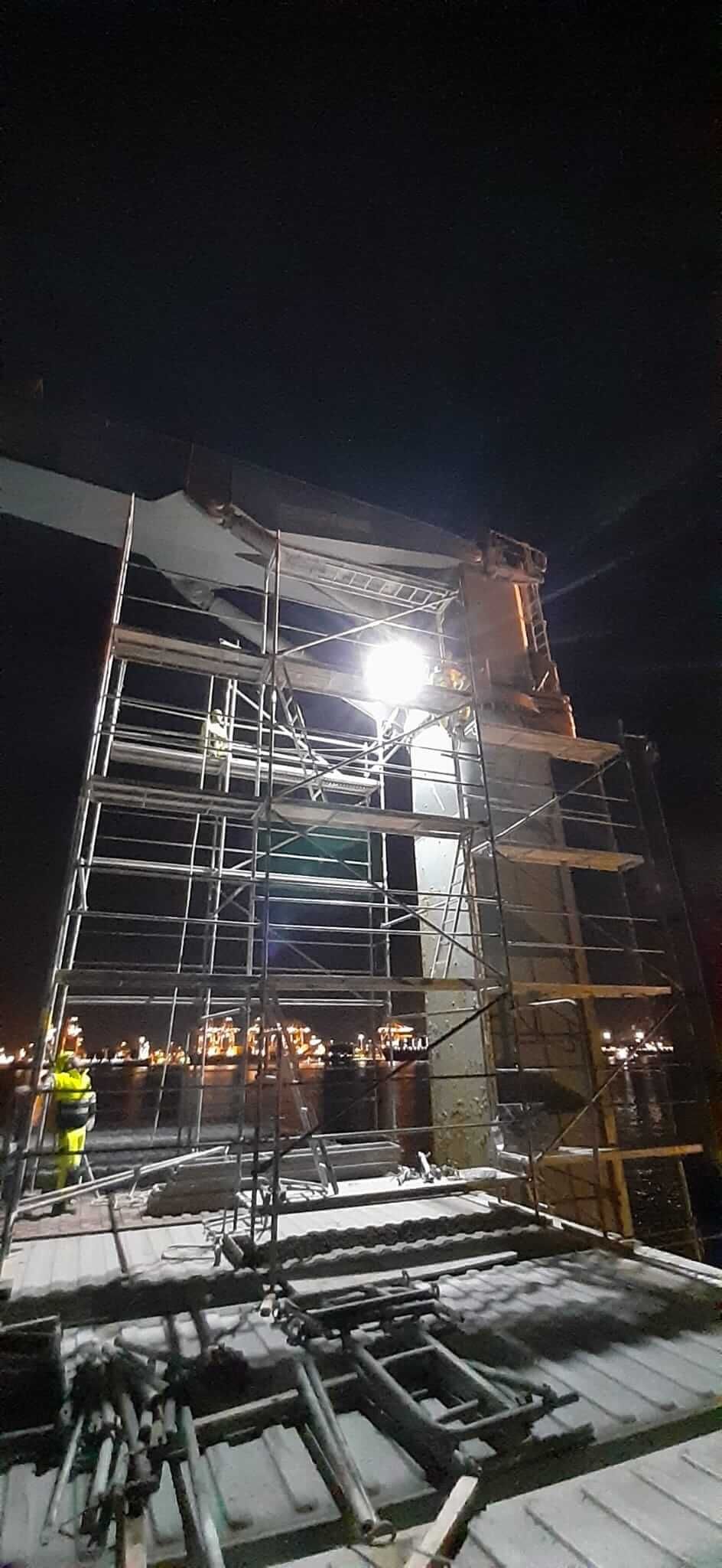 Andamios de construcción de noche, con un trabajador visible. Un foco brillante ilumina la estructura. Luces de la ciudad al fondo.