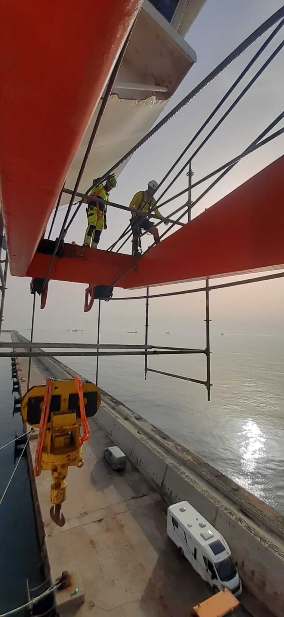 Trabajadores en un andamio sobre una estructura roja, cerca del agua. Una grúa cuelga debajo. Se ven un muelle y vehículos.