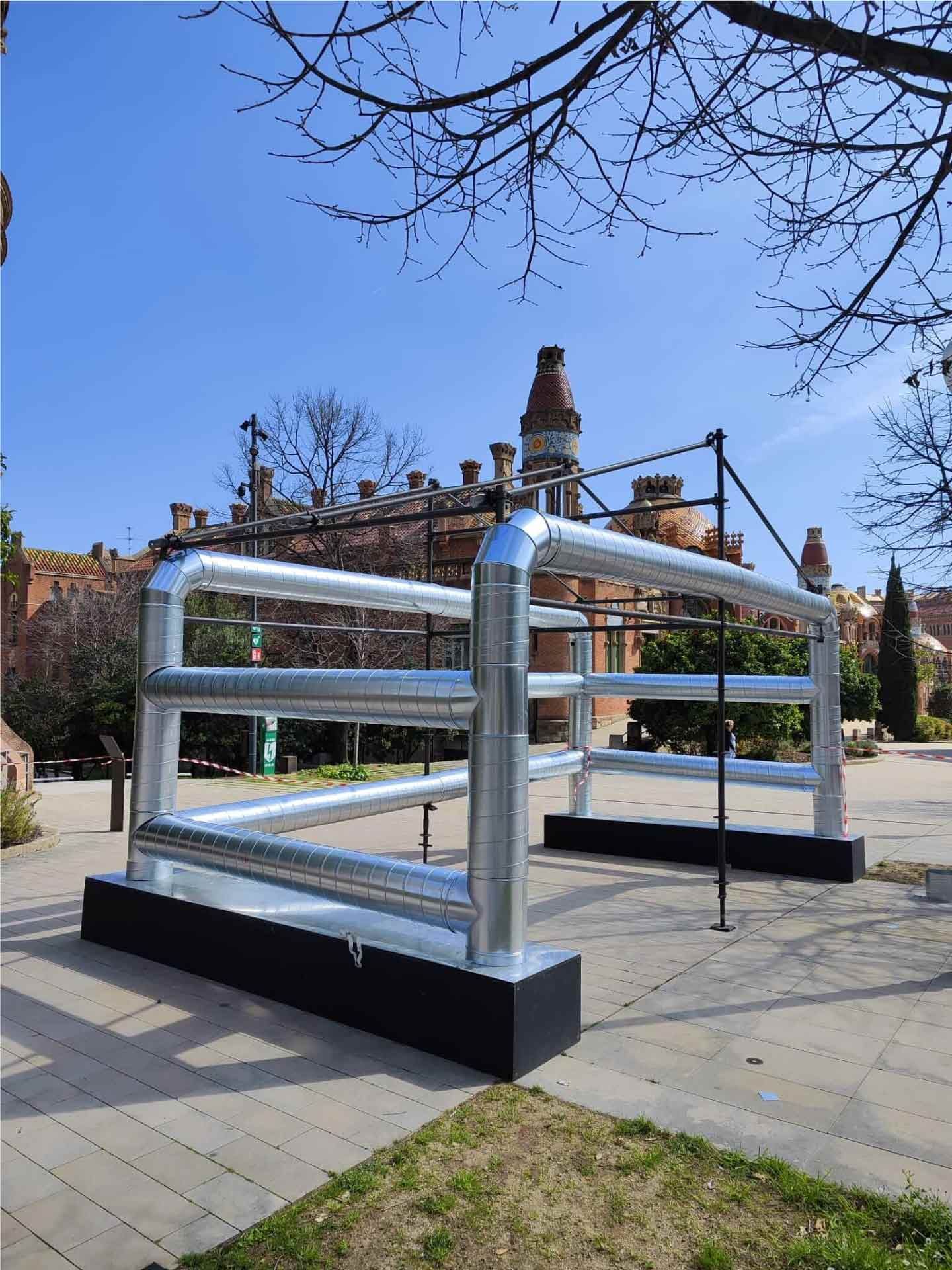 Escultura de metal plateado en un parque, rectangular con barras horizontales. Edificio histórico al fondo.