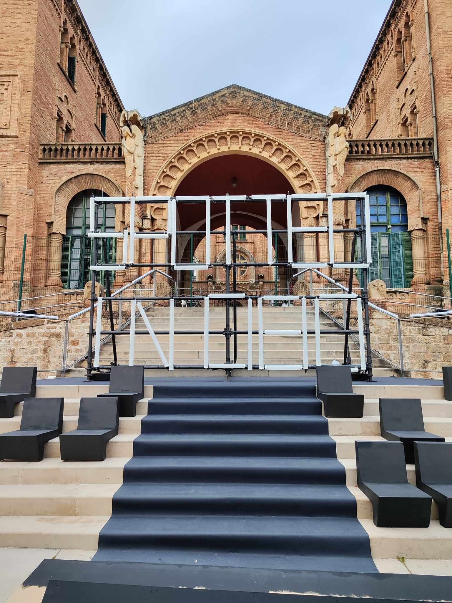 Escenario al aire libre con estructura de marco blanco, asientos negros y fondo de edificio de ladrillo.