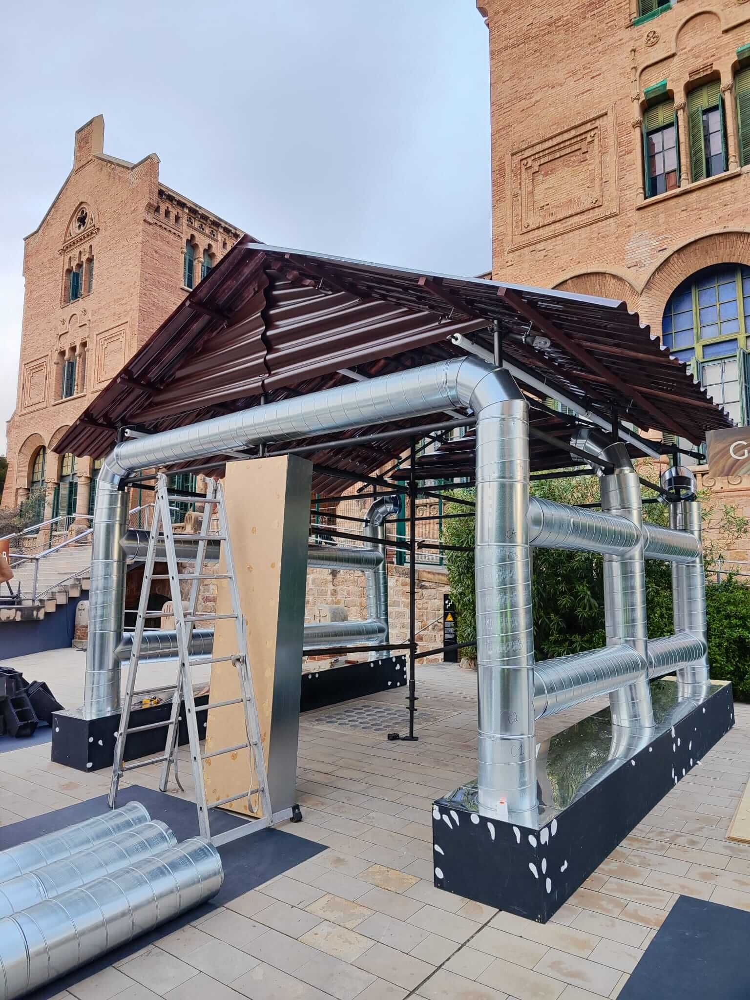 Estructura con tubos metálicos y techo marrón. Obra en construcción, edificio al fondo.