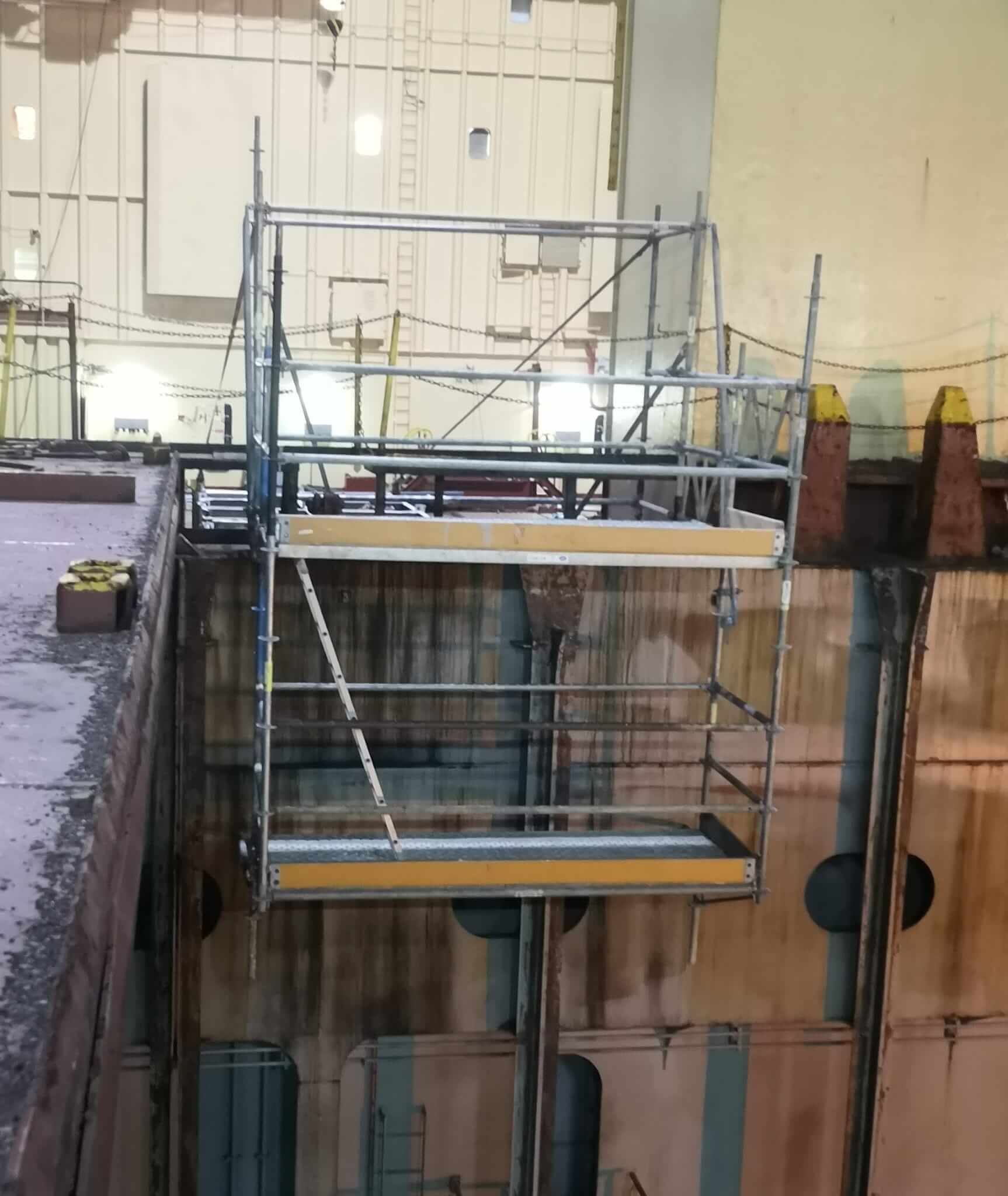 Plataforma de andamio en un entorno industrial; la plataforma se extiende sobre un desnivel. Elementos amarillos y grises.