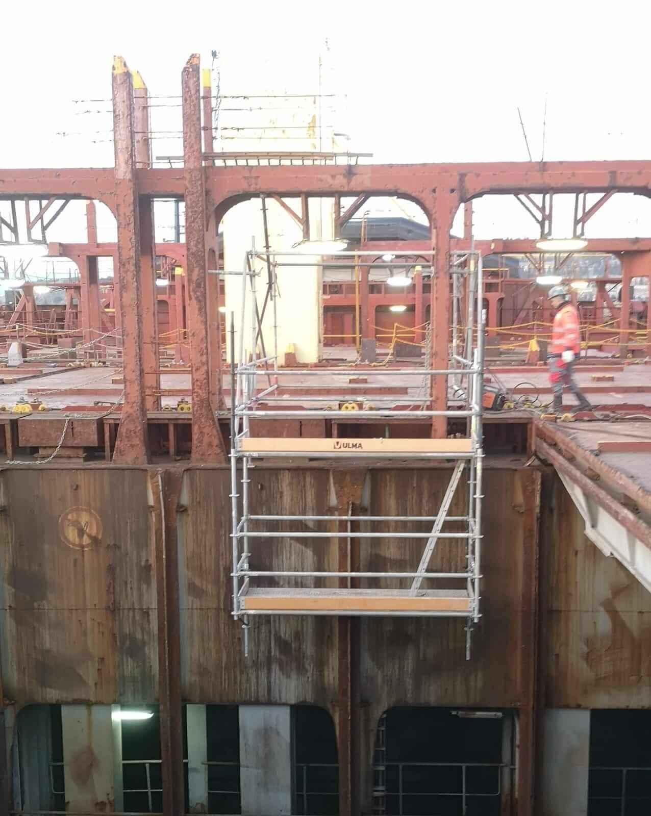 Plataforma de andamio suspendida dentro de una estructura de metal oxidada; una persona camina cerca.