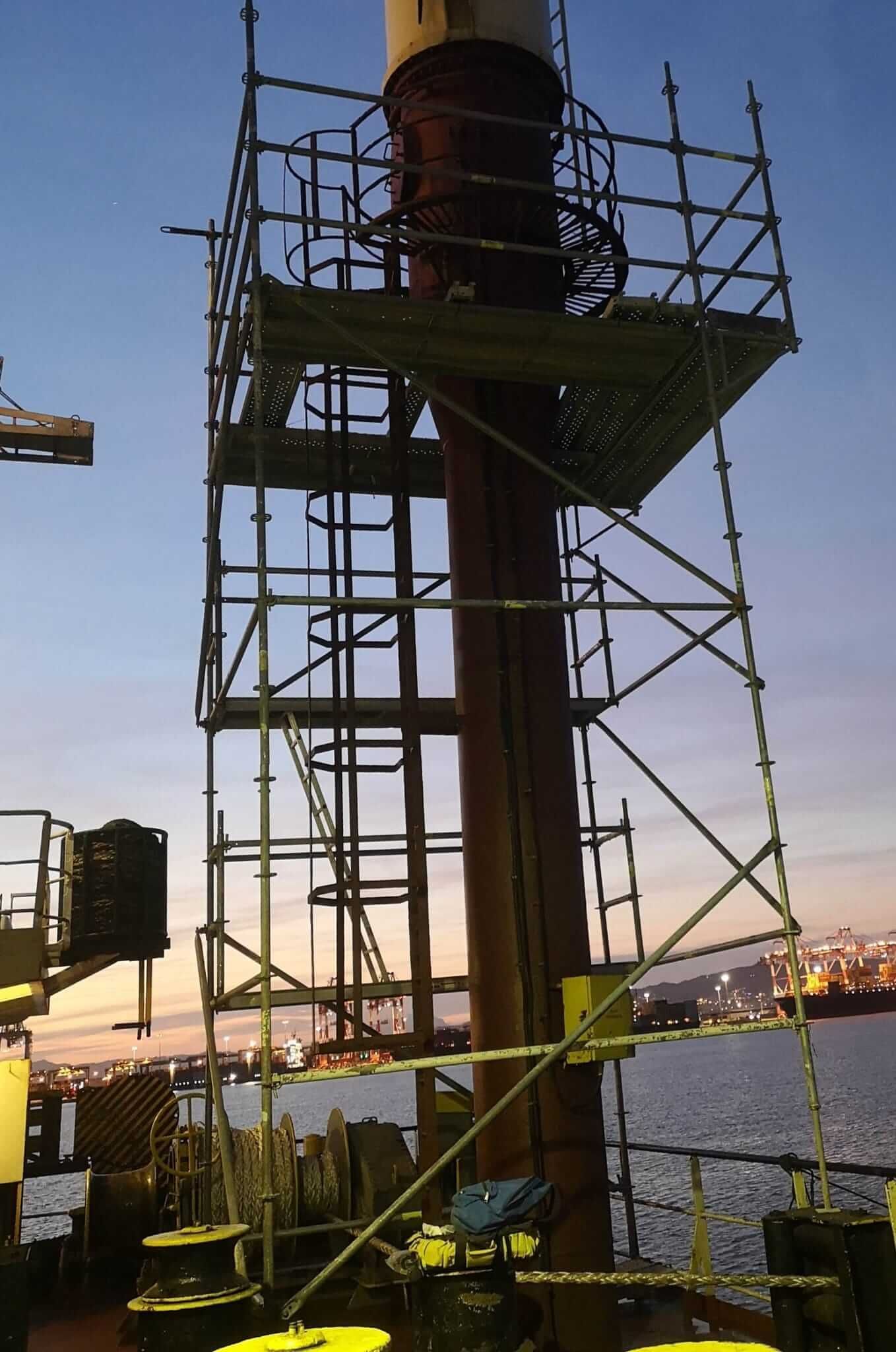 Andamios envueltos alrededor de una estructura alta y cilíndrica en la cubierta de un barco al anochecer.